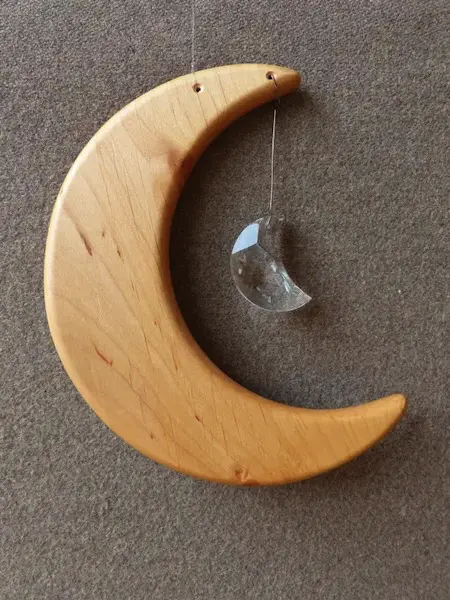 Ein Holzmobile in Form einer Mondsichel mit einem an Schnur angebrachten transparentem Schmuckstein in Mondform