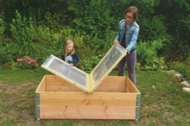 Frau mit Mädchen beim Abdecken eines Rahmenbeetes mit einer klappbaren Plexiglas-Abdeckung im Garten